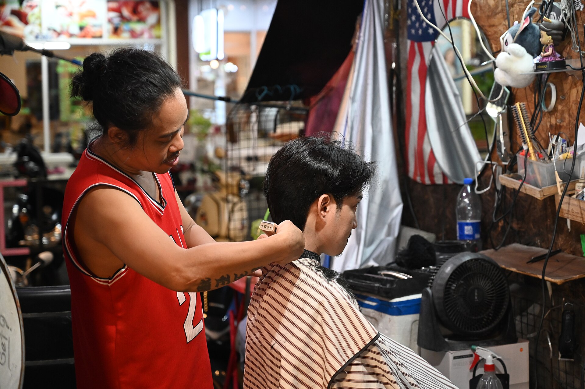 DZ6 0589 A barber carefully trims a customers hair inside a cozy cluttered shop adorned with an American flag and variou